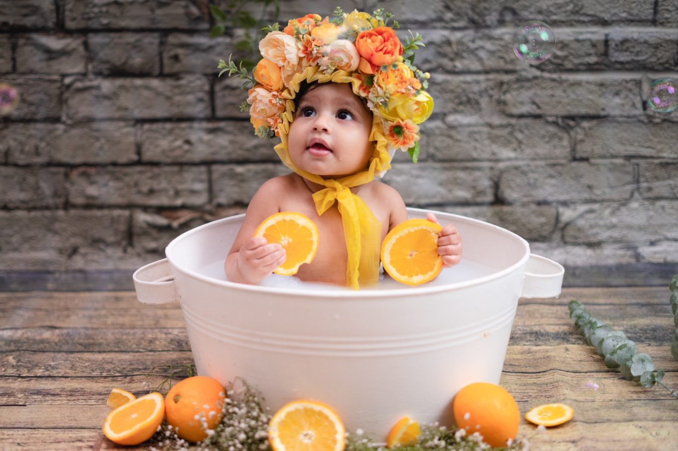 Baby in a floral crown sitting in a white tub, holding orange slices surrounded by oranges.