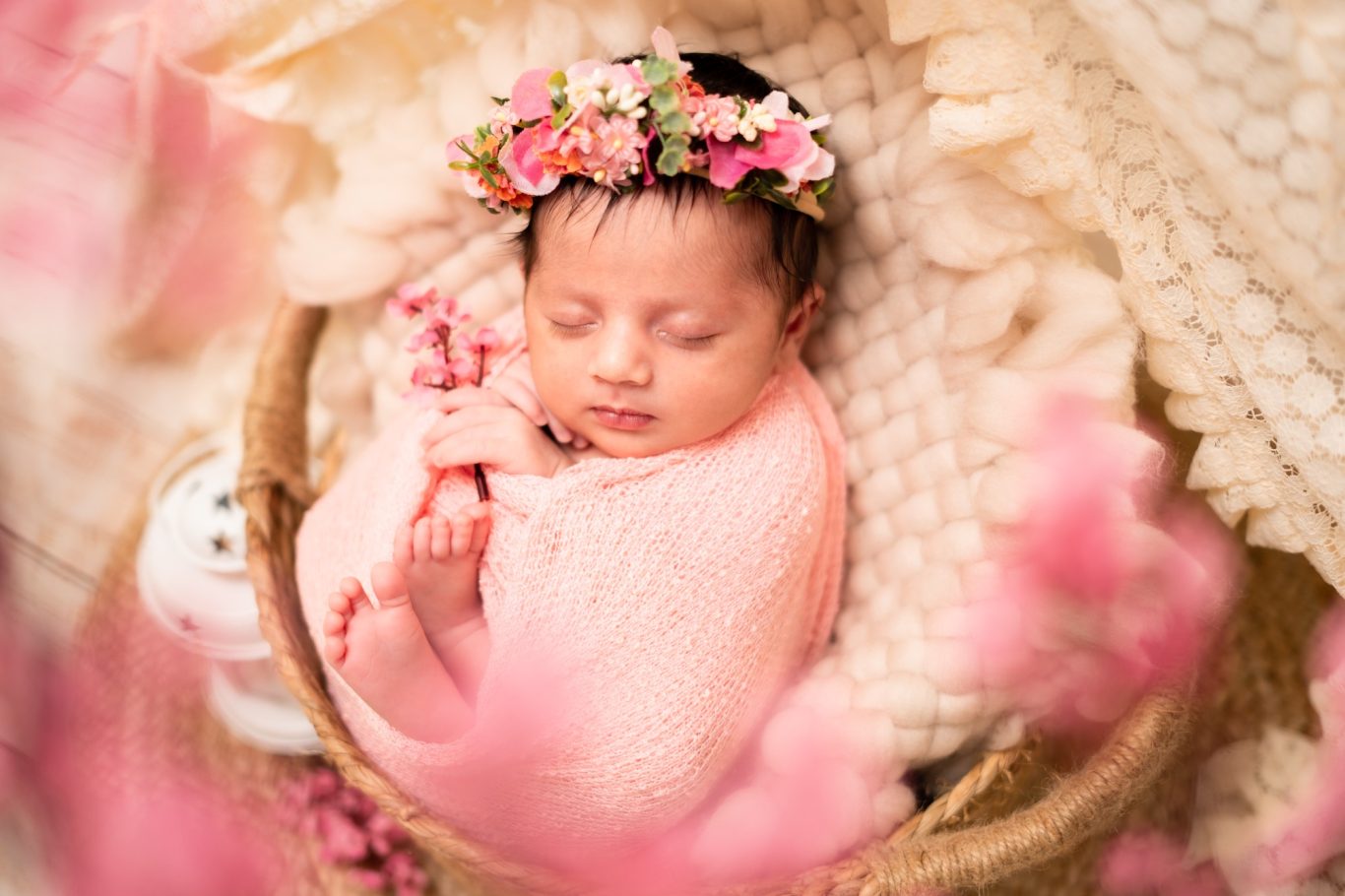 Newborn baby swaddled in pink, peacefully sleeping in a woven basket with flowers.